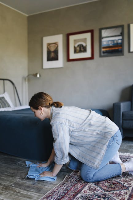 A woman with brown hair tied back, dressed in a light blue striped shirt and jeans, is kneeling on a patterned rug while cleaning a wooden floor with a blue cloth. She appears focused on surface clean