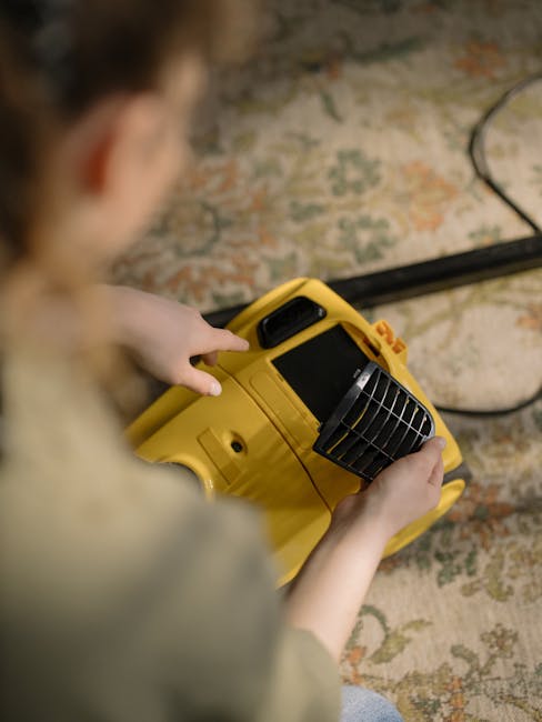 A person kneeling on a patterned rug in a residential setting, using a yellow and black portable vacuum cleaner for surface cleaning. The individual is wearing a beige jacket and blue jeans, and is ad
